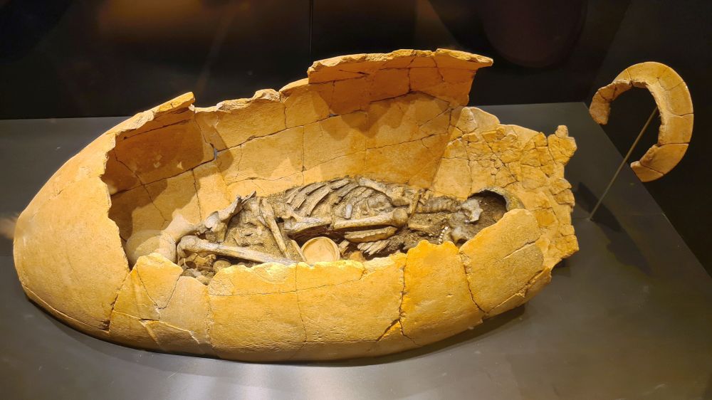 A broken clay pot, oval shaped, lying on its side. Partially visible in the clay pot, a human skeleton in fetal position.