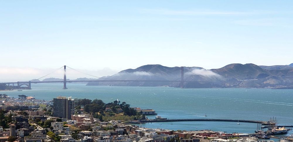 A view of Golden Gate Bridge in the distance, with its two tall towers. In this photo there's a thin layer or cloud stretched above the bridge.