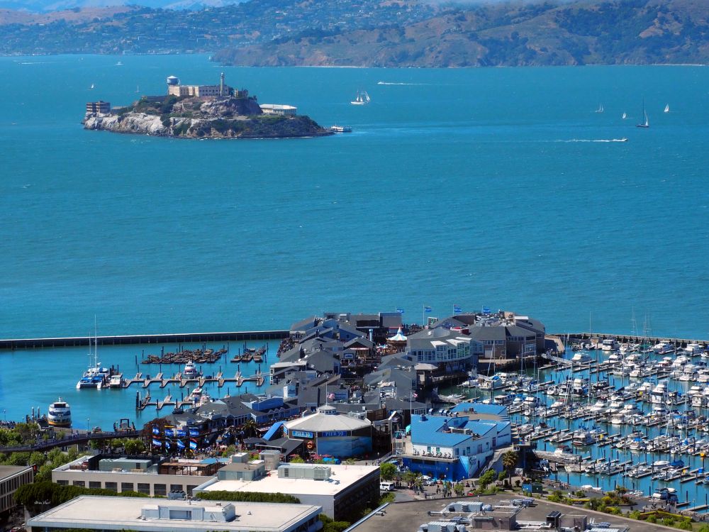 view of Pier 39, covered with buildings. On the left and the right of it are harbors. The one on the left is mostly empty while the one on the right is crowded with pleasure boats. Open water beyond that, with Alcatraz Island at the top of the picture.