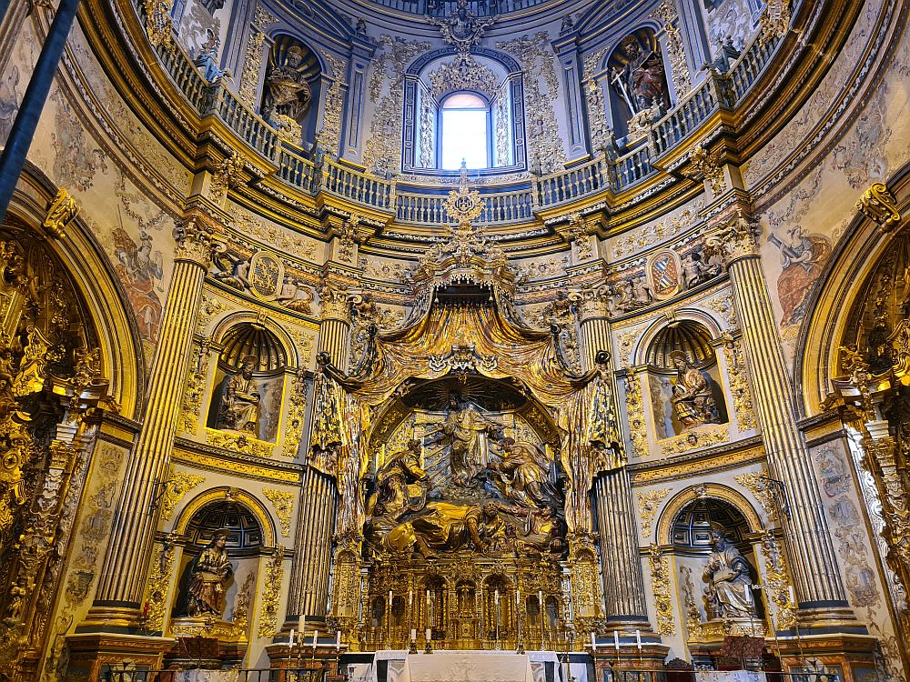 An extremely ornate altar, with full-sized statues in what appears to be mourning, and saints, possibly, on either side. Every inch of the walls going all the way up to an upper row of windows is decorated, much of it in gold.