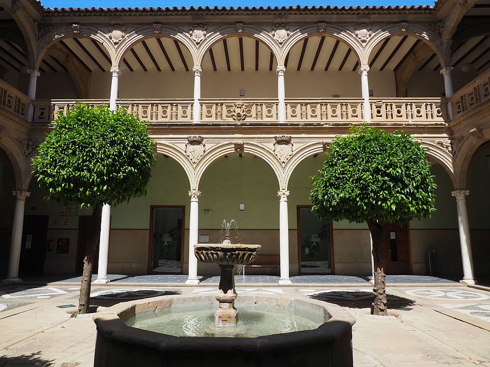 Courtyard: fountain in the center, portico along the sides, with arches supported by columns on both the ground and upper floors. A tree on either side of the fountain.