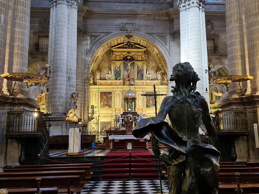 Foreground, the back of a human statue; background, huge Corinthian column clusters on either side of a central altar set into an alcove. The altar is gold-covered, with ornate carvings and many niches with statues in them.