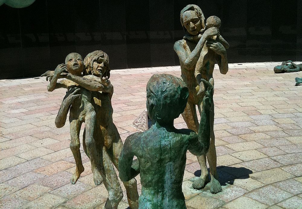 A group of statues of people: a child holds a baby, a mother holding a smaller baby, and a separate child. All are naked and made of metal with a green patina.