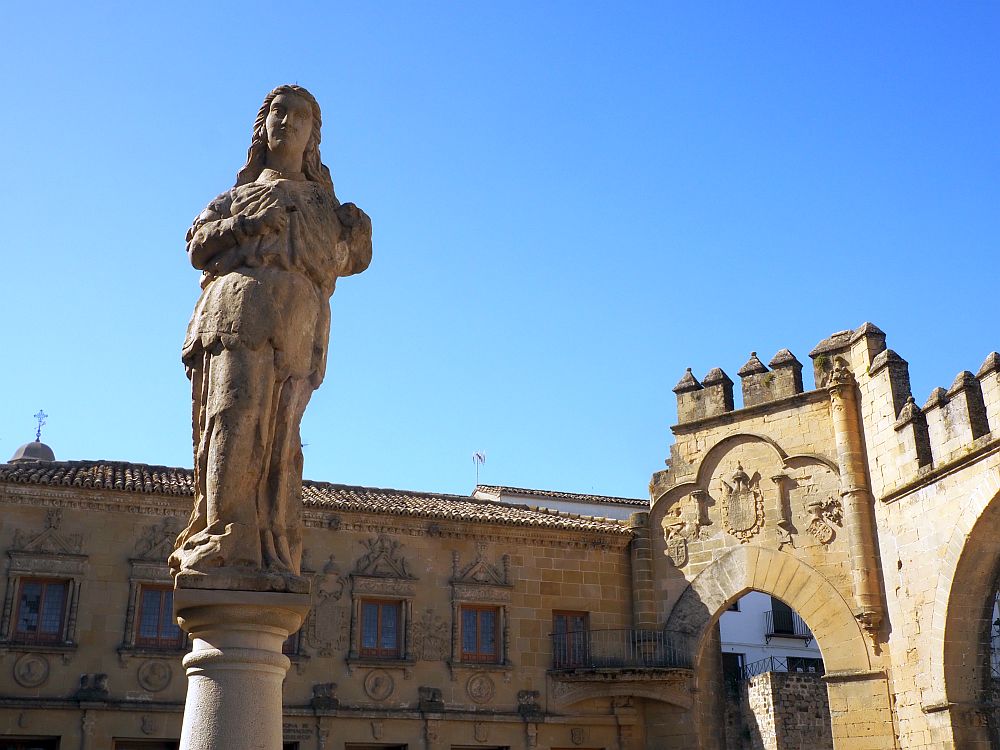 The statue is of a woman in flowing robes, presumably a goddess. The gate behind has a gothic arch and some coats of arms carved above it.