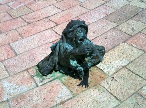 A statue of a child siting on the ground, looking up and crying as she reaches her hand out for help. Miami Holocaust Memorial.