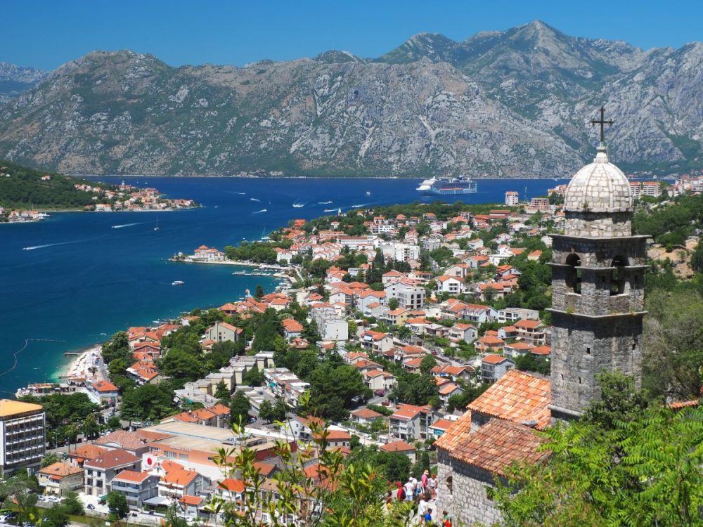 On the right, the steeple of Our Lady of Remedy. Below, the city of Kotor and beyond it, the blue water of the bay, with hills on the far side of it.