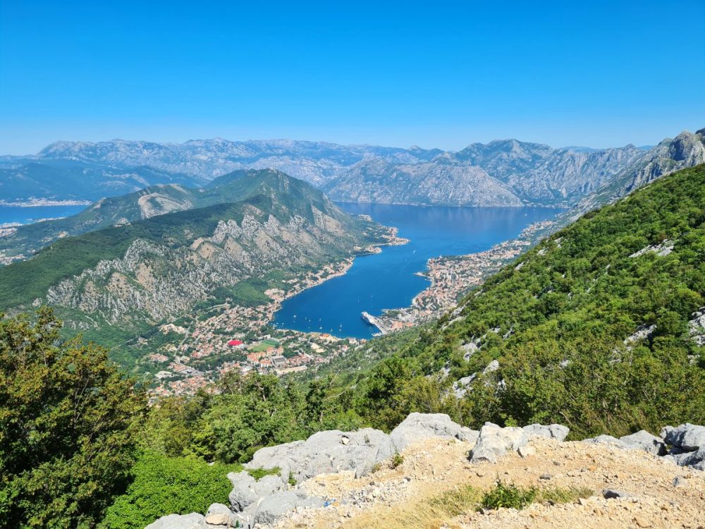 Far below, a long body of water - the Bay of Kotor - flanked by steep rocky hills with shrubby growth. Far below, around the edge of the water, the city of Kotor.