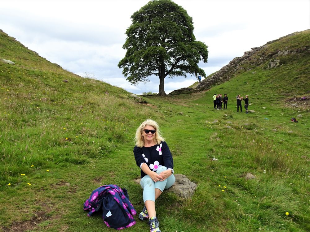 Elaine Wilson posing in front of the Sycamore Gap tree, with Hadrian's Wall running right by it.