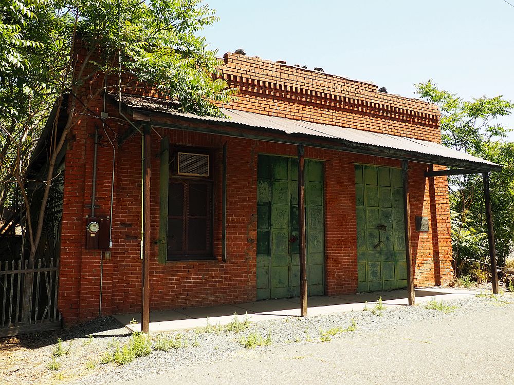 Single-story brick building, a roof shading the walk in front of it. One window and two large doors, shuttered closed with green shutters.