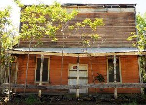 A house that looks like two stories, but the upper one has no windows: a false front. The ground floor has a door in the center and a window on either side, painted a faded yellow. A roof shades the sidewalk or porch in front.