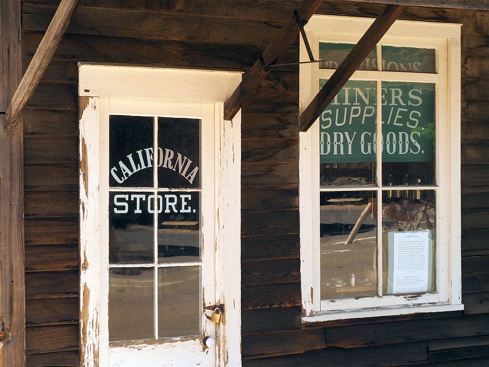 Close-up of a brown-painted storefront with a door and a window, both edged in white trim. A sign on the door reads "California Store" and a sign in the window reads "Provisions, miners supplies, dry goods."