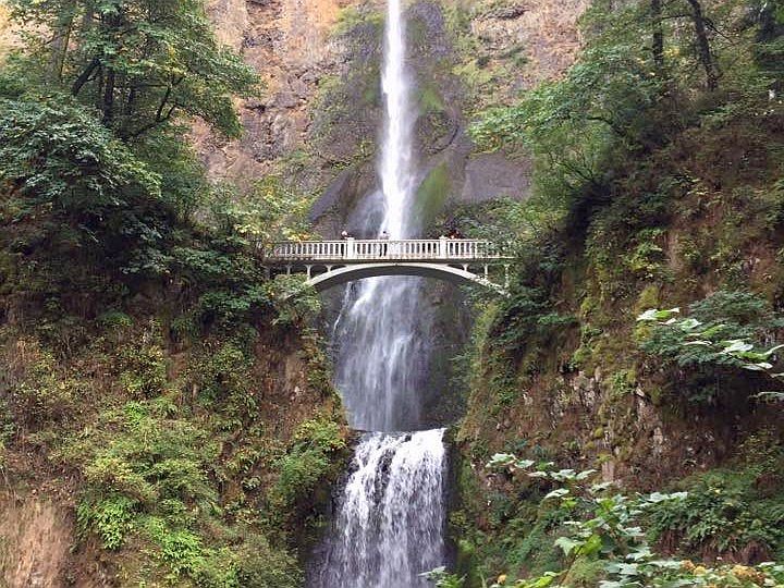 A long narrow waterfall - even longer than the photo shows, since it crosses the entire picture top to bottom. A small bridge crosses in front of it about halfway up, with people barely visible on it. 