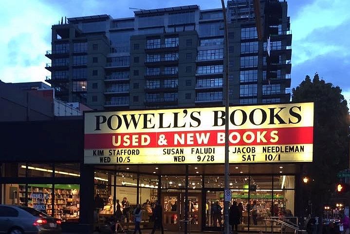 Powell's Books with a huge sign across the entire width of the building, lit up bright at night. Lights inside the store reveal bookshelves and lots of people. 