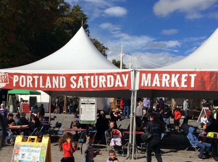 An open-sided tent with the words "Portland Saturday Market" along its edge. People under it sitting at tables, eating or just sitting.