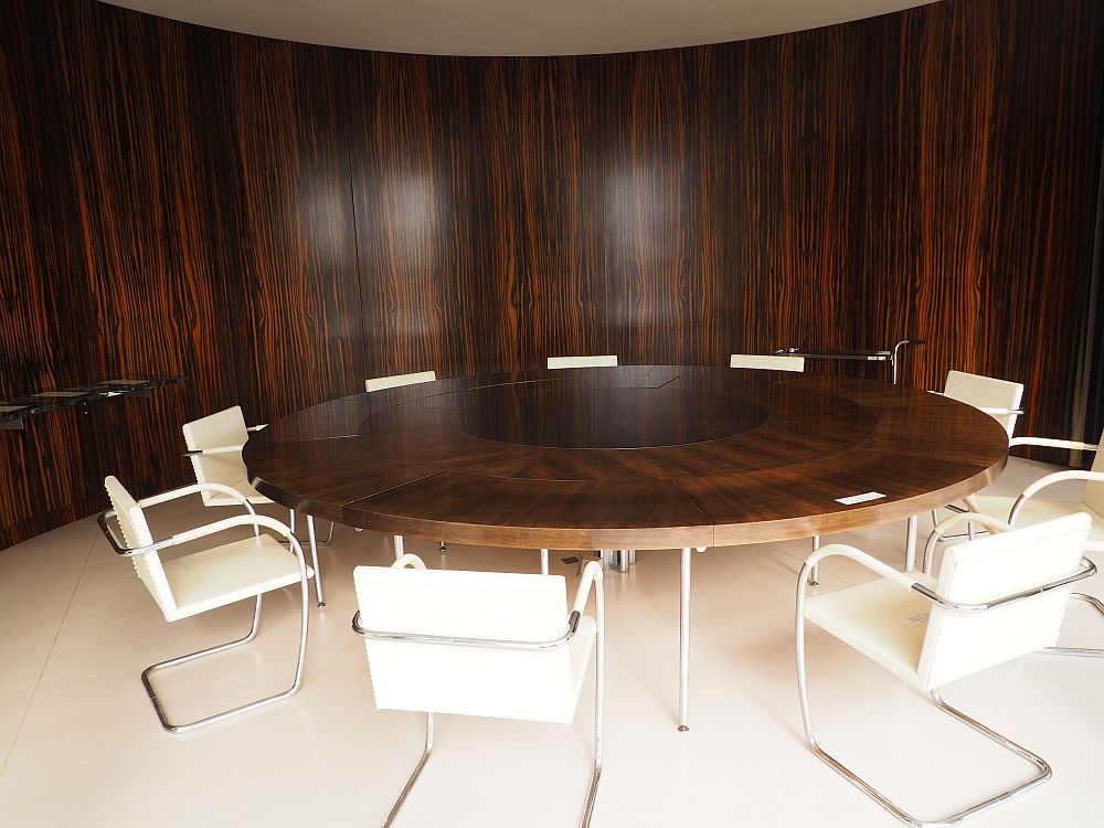 View of the dining room, with a large round dark wood table surrounded by white chairs with curved tubular frames. Behind it, a curved dark wood wall with a vertical grain.