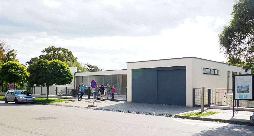 A two-car garage on the sidewalk, then, next to that the rest of the house is set a bit back from the road: white, with a wide glass front and a flat roof, very simple. 