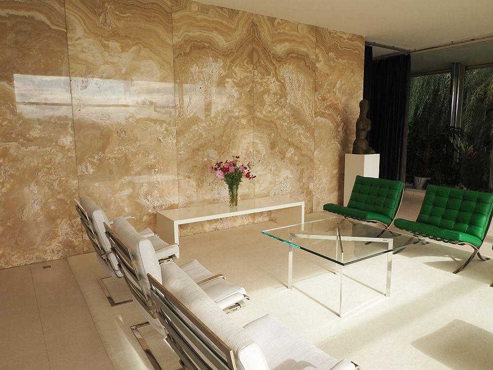 A view of the Villa Tugendhat living room space. Two rows of chairs face each other: one white and one green. The back wall is onyx: shiny with a brownish-yellow grain visible.