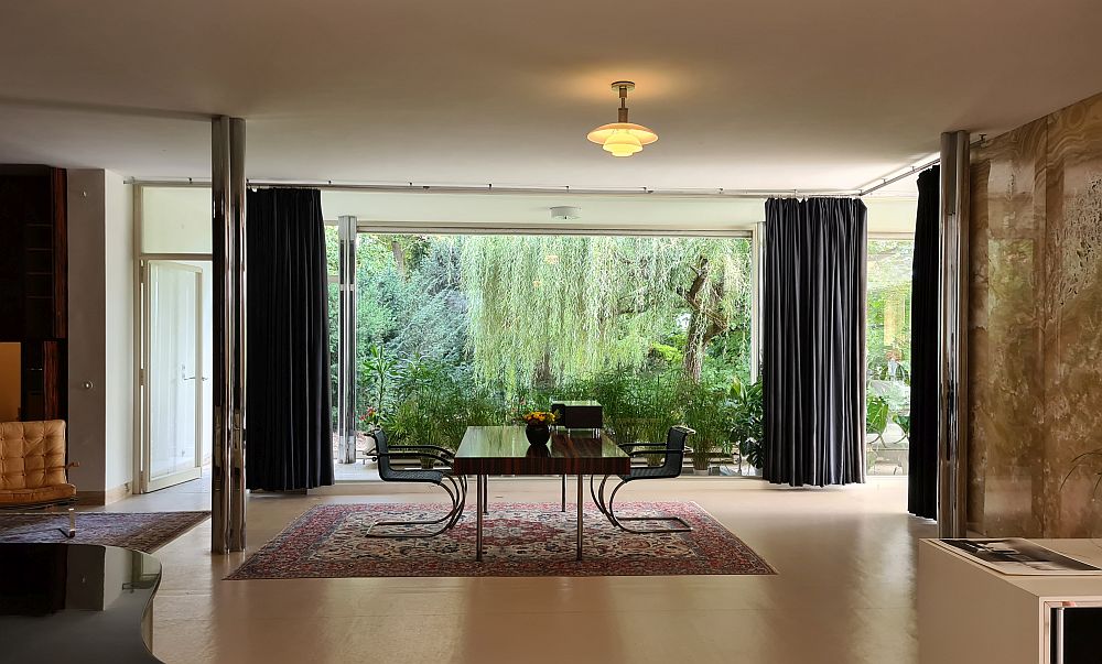 Straight ahead, a large room with a big desk in the middle, a chair on either side of it. Beyond the desk, a floor to ceiling window with potted plants and a garden visible beyond that.