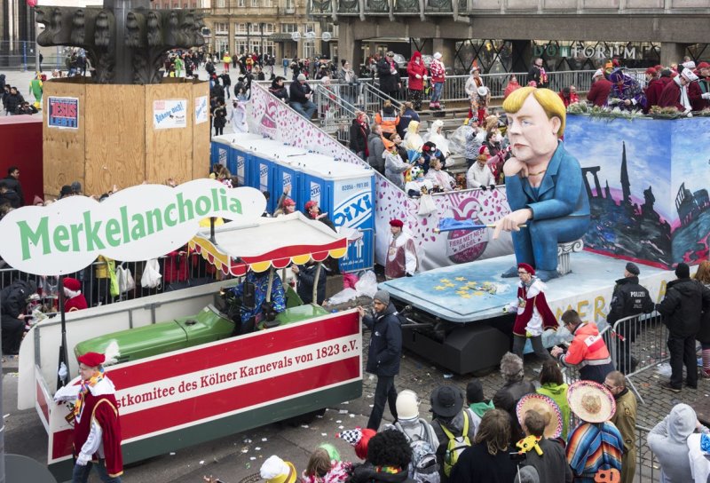 Spectators in stands along a street behind. In front, a float passes by with a sign saying "Merkelancholia" and a huge statue of Angela Merkel.