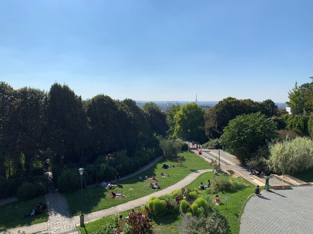 View over a green grassy area and woods, with, in the distance, a view of Paris including the Eiffel Tower.