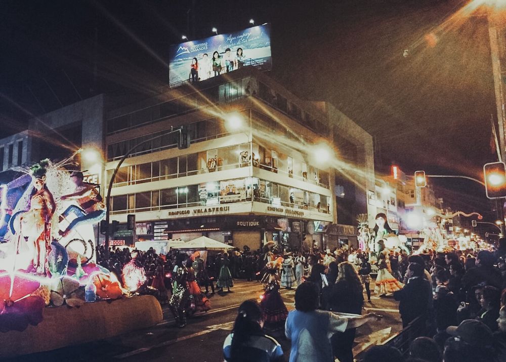 A street corner at night: crowds of people, a brightly-lighted float on the left, a row of floats visible approaching down an avenue on the right.