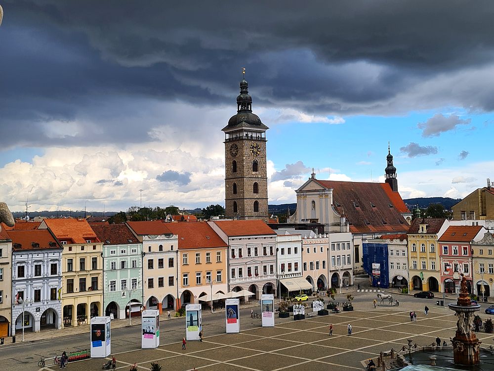 Ceske Budejovice central plaza seen from slightly above: a row of pretty pastel buildings, with a tall stone tower behind them.