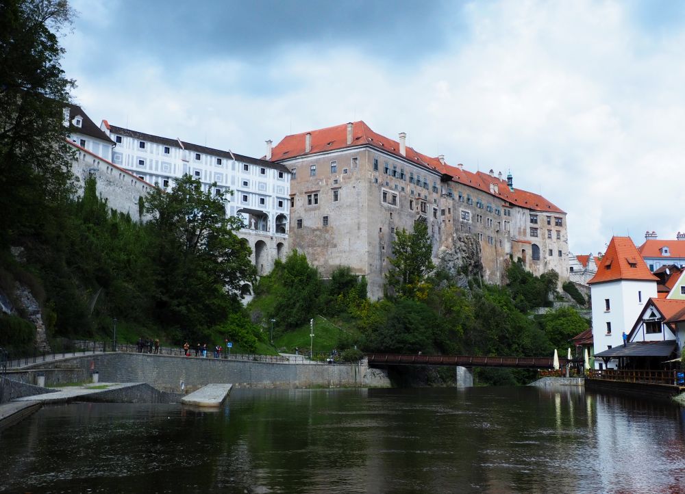 Cesky Krumlov castle rises high above the river, its multi-story bridge partly visible. On the river, two parallel walls ahead and a bit to the left channel the water into a gentle slope.