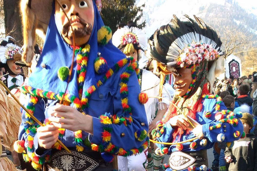 People in blue outfits with a wide belt and strings of garlands along the sleeves and collars. Each has a mask on and a tall feathered and flowered headdress.