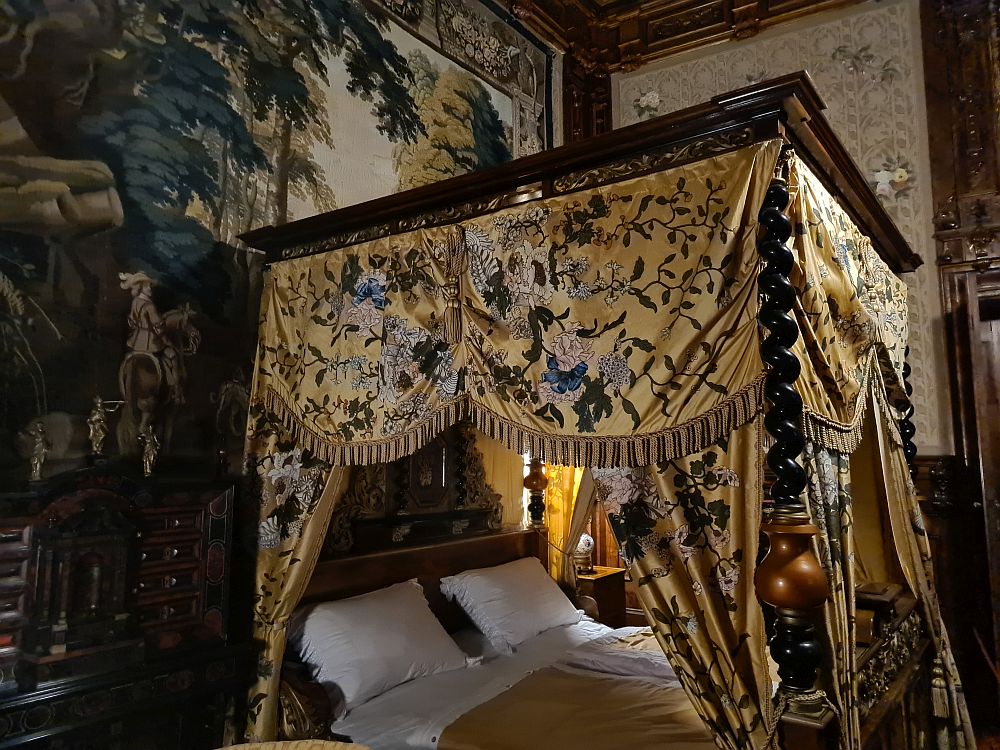 A four-poster bed with a cloth covered in detailed embroidery. On the wall behind, a massive tapestry.