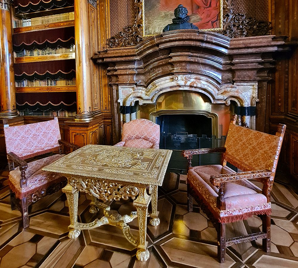 A gold table with three chairs around it in front of a marble fireplace that incorporates at least 2 different types of marble.