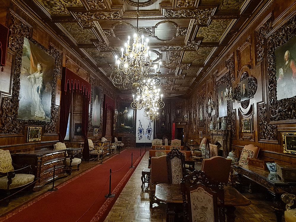 Looking down a long room, chandeliers down the middle of an ornately-carved ceiling, groupings of furniture, ornately-panelled walls framing large portraits.