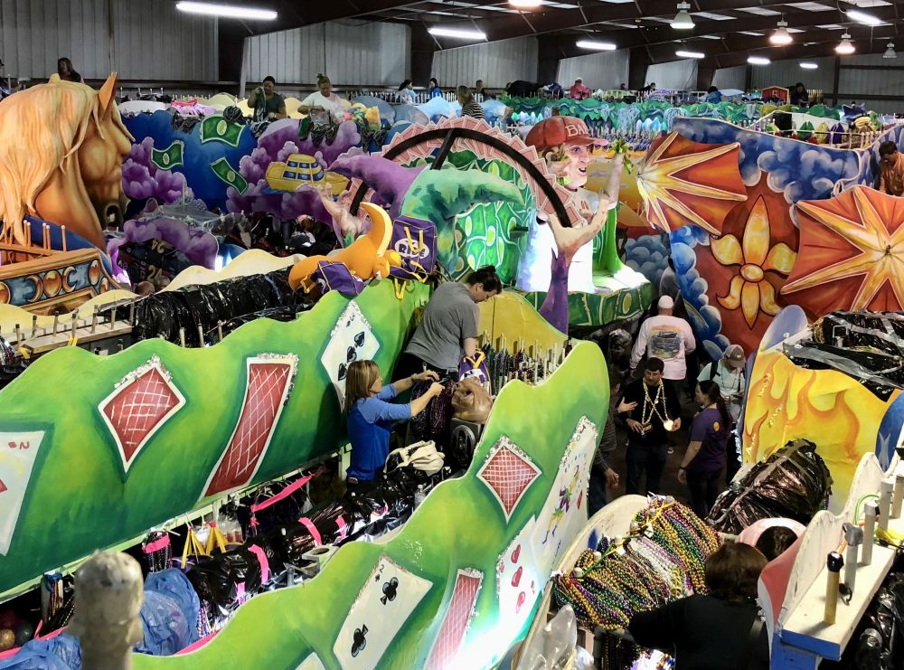 A very busy scene inside a large warehouse-sized hall crammed with floats in bright colors. On the nearer ones people can be seen, working on the floats or speaking to each other in the small space between floats.