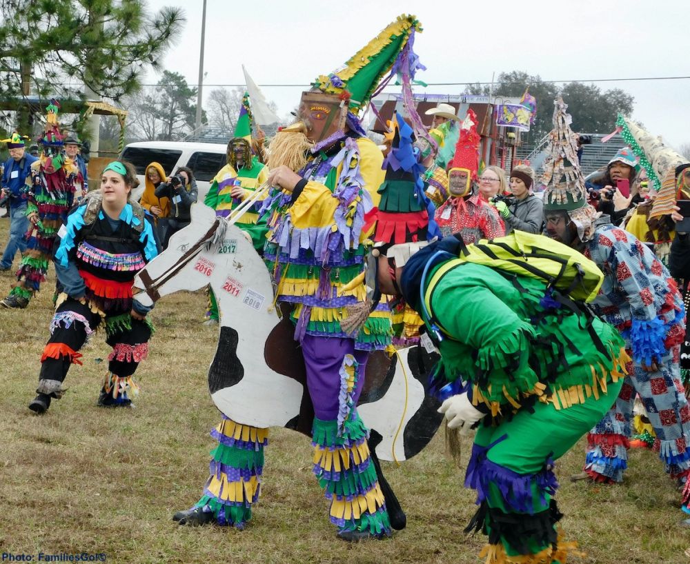 A person, wearing a tall hat, a mask with a pointy nose, and a frilly costume in green and yellow, stands straddling a cutout of a horse, painted in white and brown, looking like he's going to ride it, but with his feet on the ground. Beside him is another person preparing to run in a similar costume, but without the horse cutout..