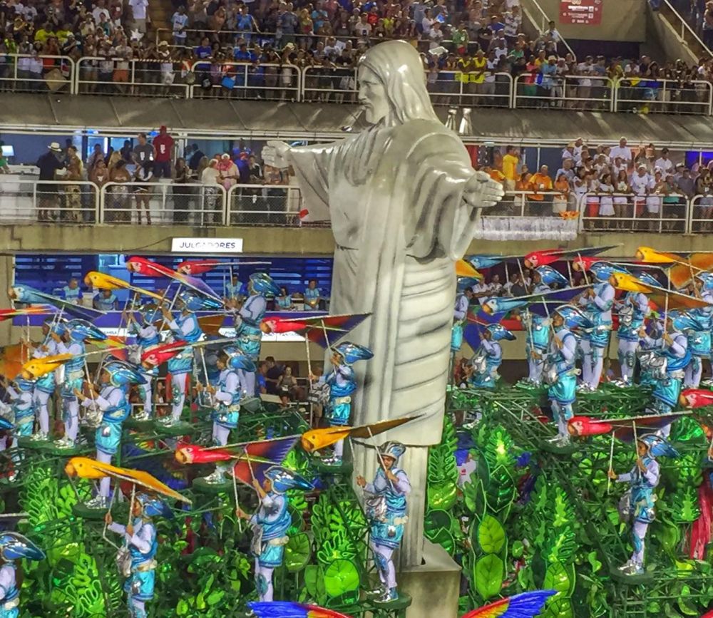 Dancers in blue stand on a framework meant to resemble a forest, each holding a large kite that looks like a bird. A huge statue looms above them that looks like the statue of Christ the Redeemer that stands on a hill above Rio de Janeiro.