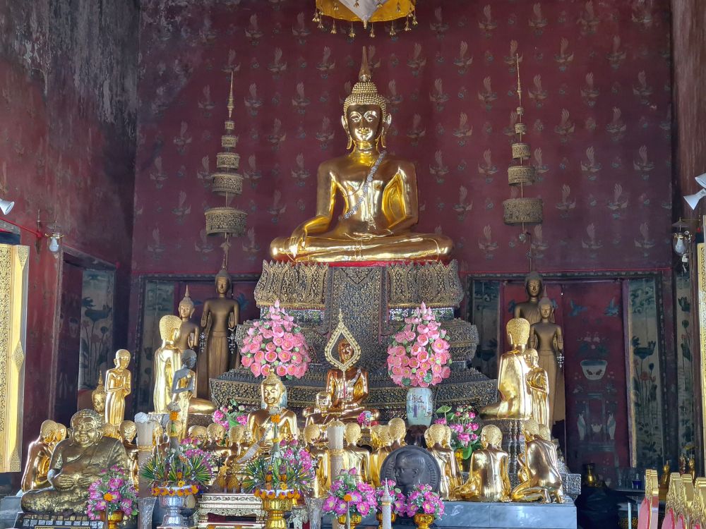 A gold Buddha statue sitting quite high on a pedestal in Subduing Mara pose. Below it, an array of other gold-colored statues, some standing, some sitting, mostly facing the large Buddha in the center.