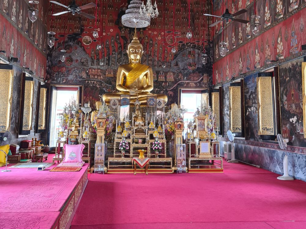 A view down the inside of a temple, with the usual gold Buddha up high on a pedestal at the end - this time in meditation pose. The walls are painted with frescos. The floor is covered in reddish-pink carpet, and the left-side is raised about a food higher than the rest.