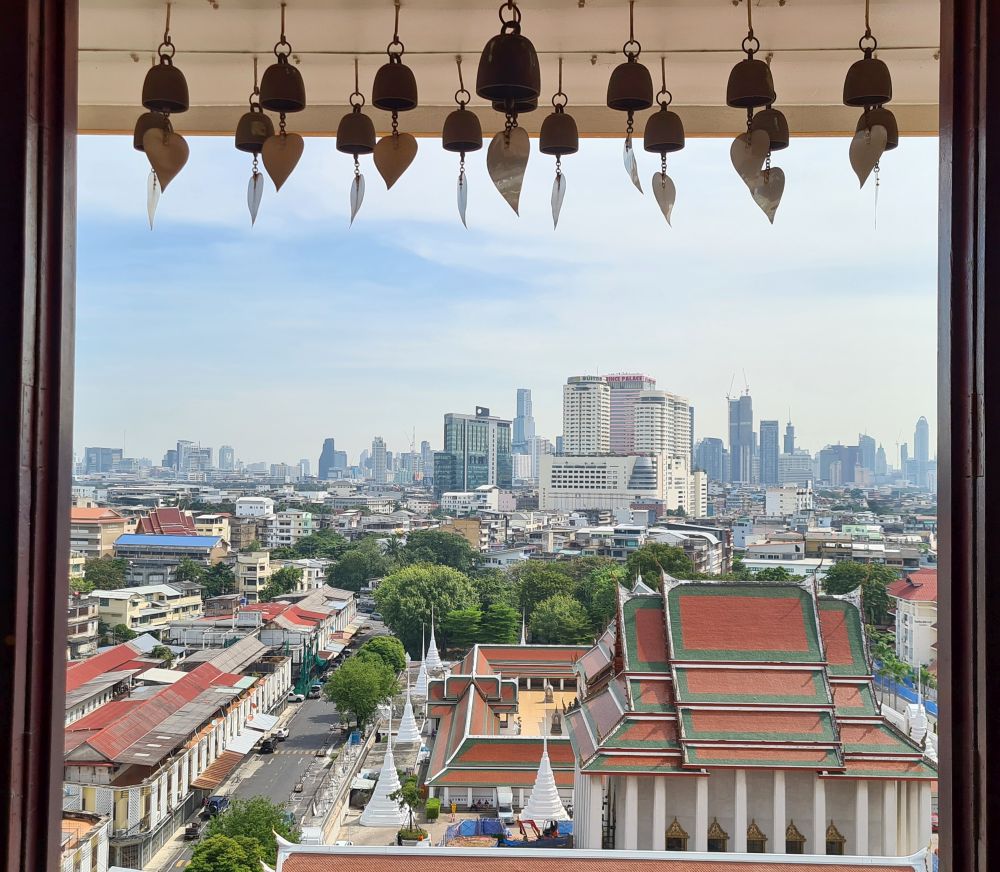 Looking through a window with a row of small bells hanging from its top, looking over a termple in the foreground with red and green roofs, and the modern city with skyscrapers in the background.