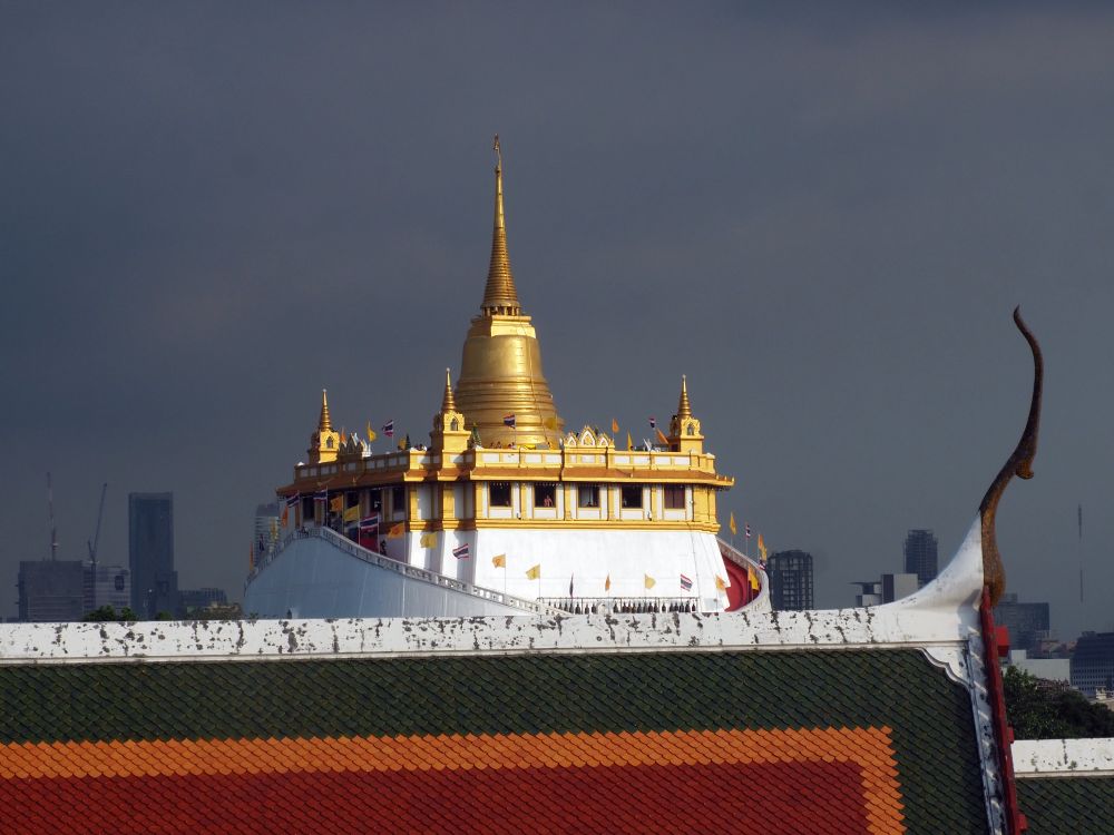 The crest of a temple roof in the foreground, Golden mount in the background: white walls and a gold stupa rising in the middle of the roof.