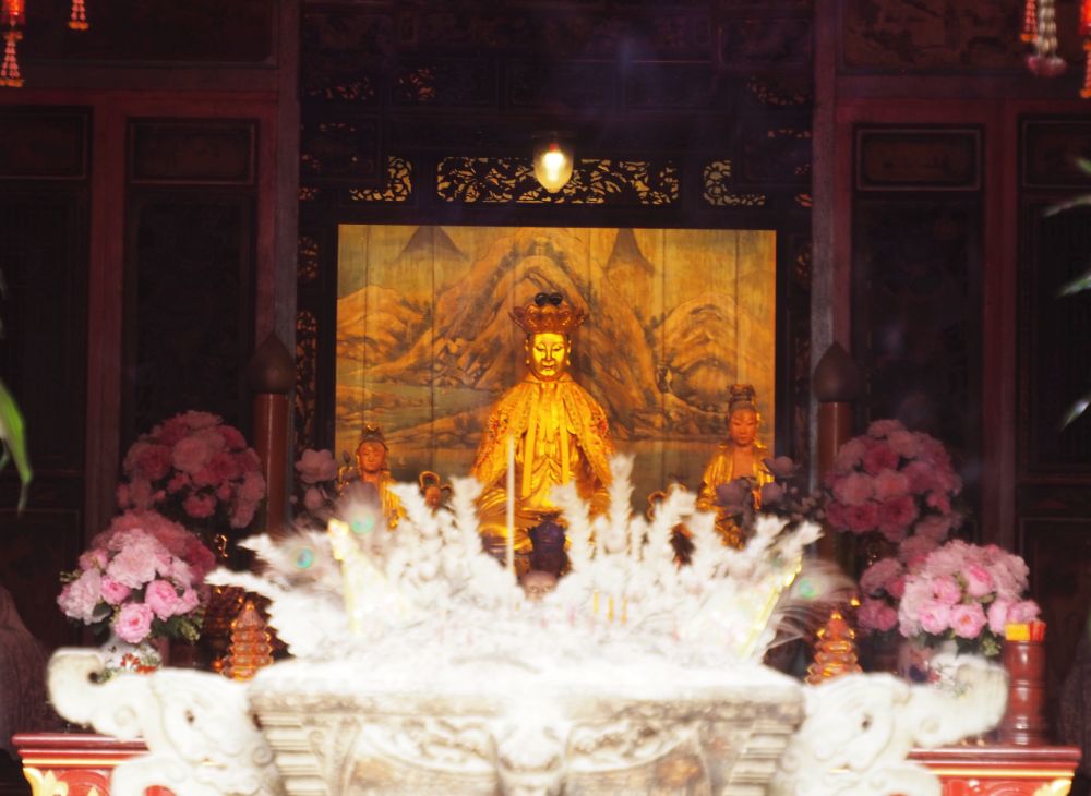 The image is gold and sitting, with a squarish hat. In front, blurry, is an altar with what may be flowers and/or feathers.