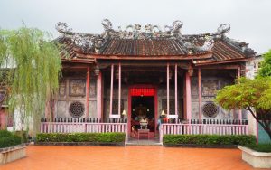 Traditional single-story Chinese temple with a central entrance and typical Chinese roof lines. Ornate carving along the peak of the roof and some faded painting on the exterior walls.