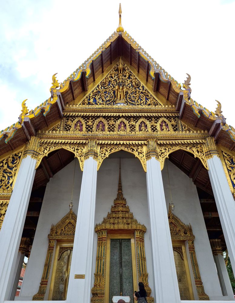 Looking up at the ornate entrance. Four white pillars hold up the gable which is decorated in gold, with rows of buddhas across above the entrance. Behind the pillars are 3 doors to the temple, all closed, and all crowned by an ornate gold decoration.