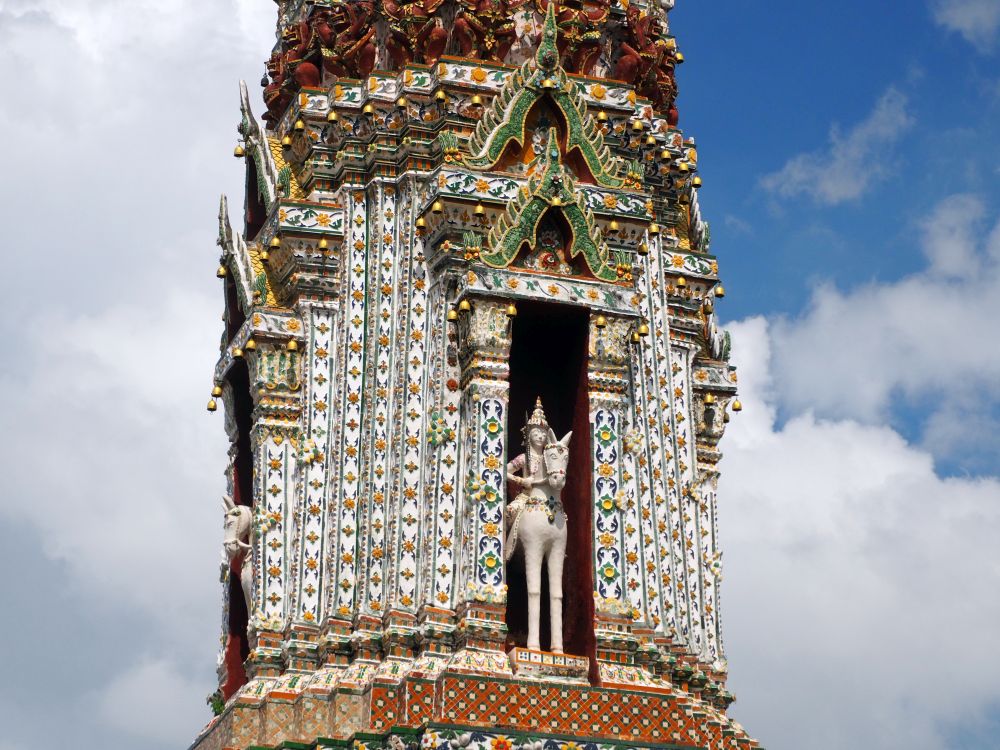 Looking at a middle level of a tall stupa; it has a tall narrow niche in it where a horseman sits on a tall narrow horse. The stupa is busily decorated with ceramic tile.