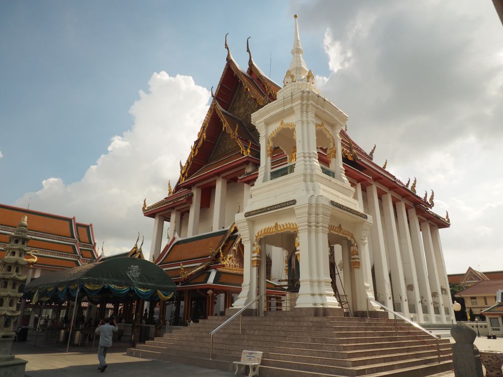 A very large temple, with high square pillars holding up a mostly red roof, decorated around the edges in gold. In the foreground, a two-story white structure that holds a bell. 