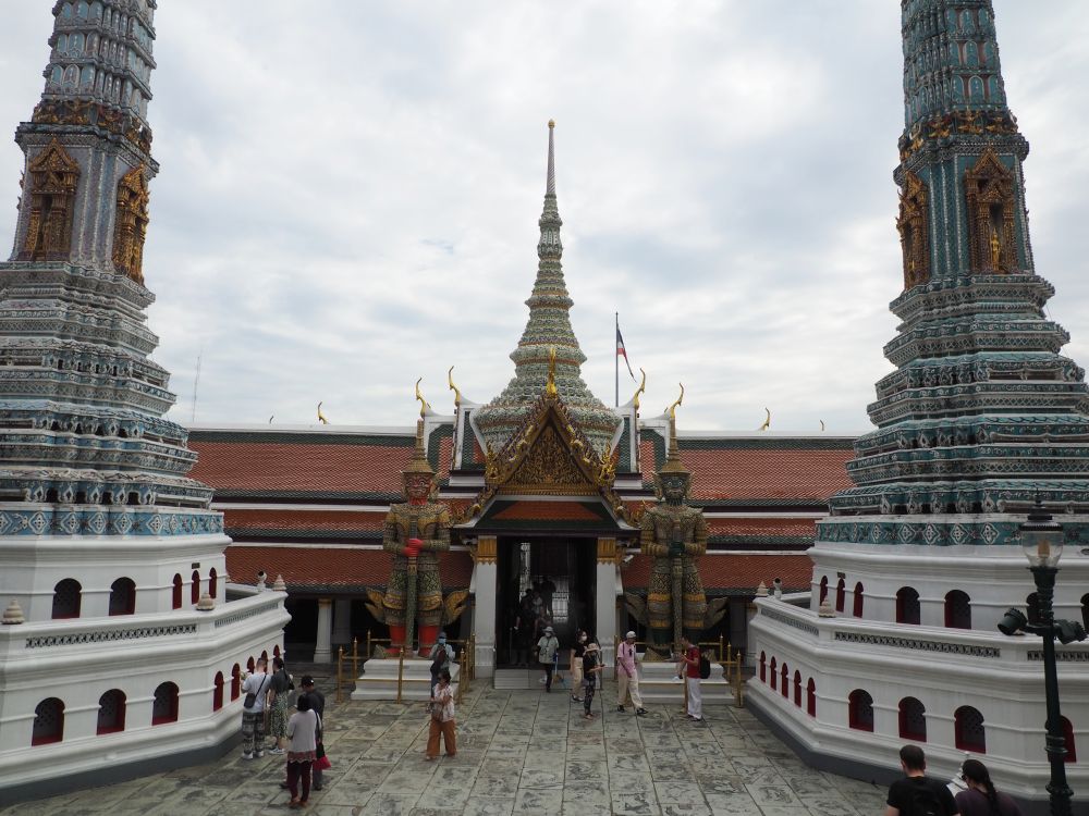 A stupa on either side, and entrance in the center of the photo with a yaksha on either side of it. Above the entrance is another stupa. The stupas are all highly decorated with tile. A few tourists walk on the space between the stupa and illustrate that the yaksha are about 3x a human's height.