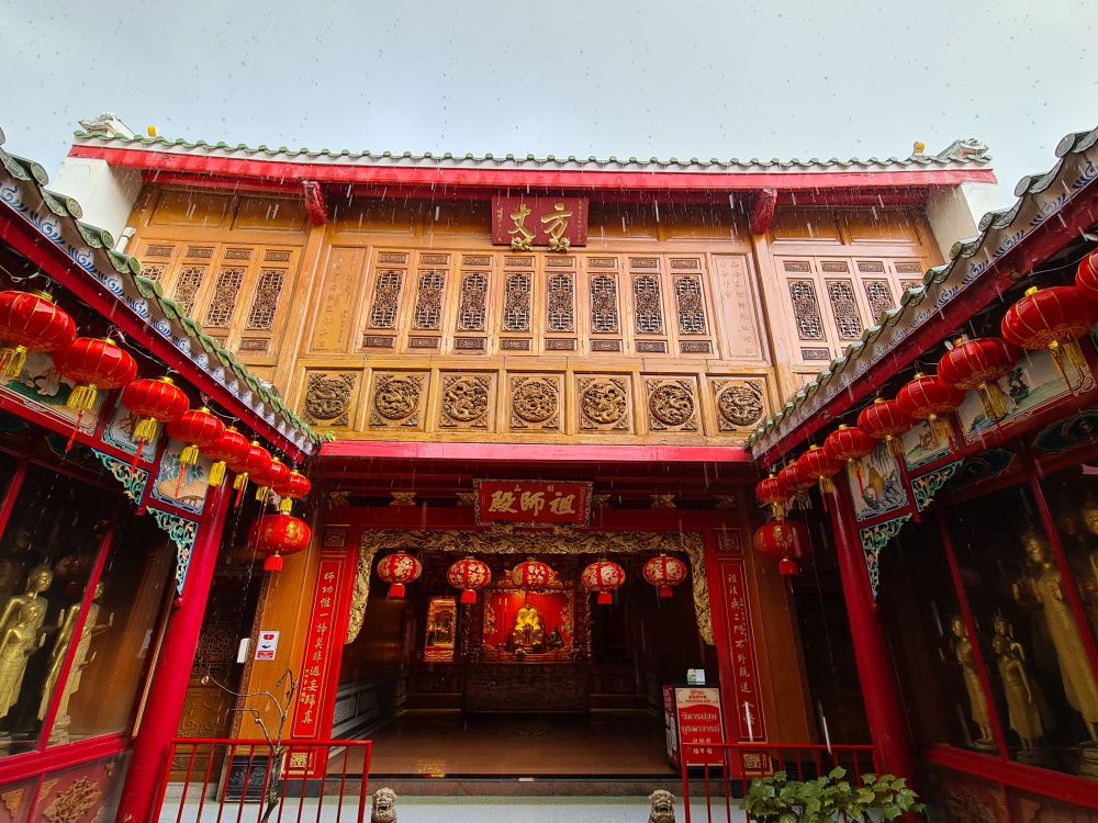 Two single story rooms on left and right, where you can see just a bit of the glassed-in display of golden buddhas. Ahead, an open-sided room with a shrine and above that, on the upper floor, a beautifully-carved wooden facade under a red-edged roof.