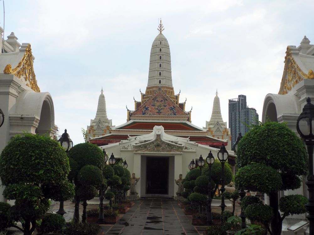 Straight ahead, a temple with decorative gable and red roofs behind that, topped with a large white stupa. ON either side of the photo, part of the two side stupas are visible, also white with gold details.