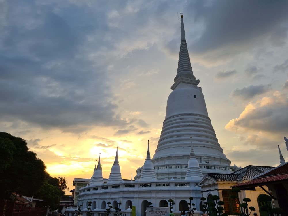 A very large white stupa with a wall around its bottom that has many smaller stupas along it. Sun setting in the background.