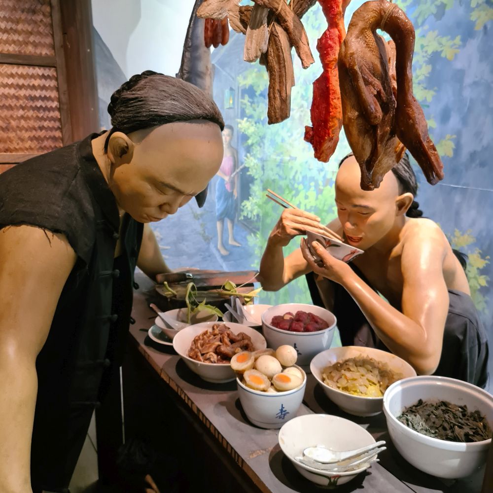 A very realistic waxwork man stands behind a counter with various bowls of food arrayed on it. Another man sits on a chair on his haunches, wearing no shirt, and eating from a bowl with chopsticks.