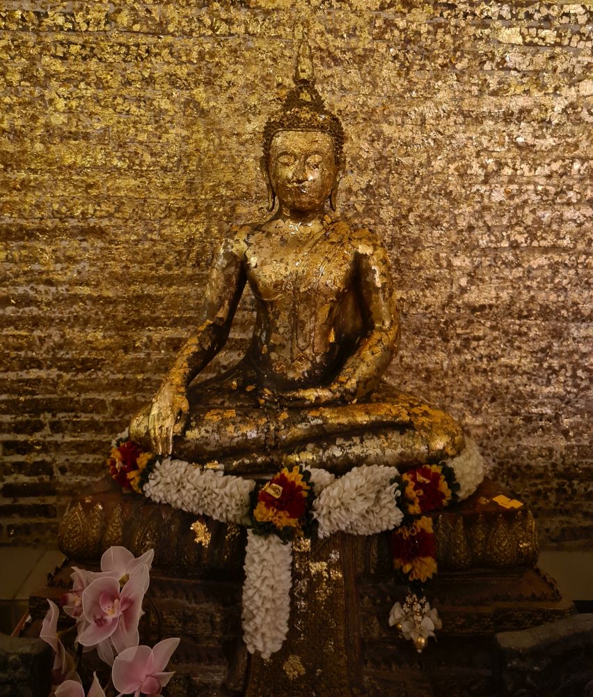 A Buddha image in Subduing Mara pose, covered head to tow with bits of gold leaf, some of which are peeling off, creating a textured effect. The brick wall behind it is also coverd with bits of gold leaf.