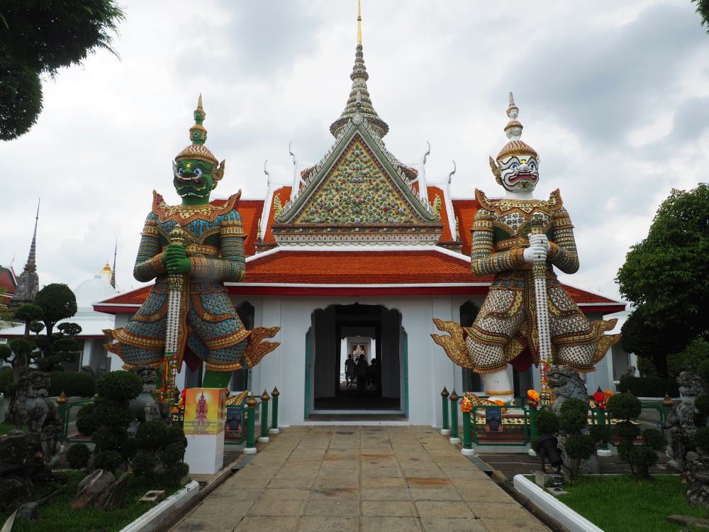 The entrance is in a traditional thai style, with a steep peak and detailed decorations under the gable, a plain red roof otherwise. On either side of the square entrnace is a yaksha: large humanoid characters in colorful outfits but with animal-like faces, clutching swords and wearing tall helmets.